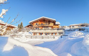 En stor chalet i trä med flera våningar och balkonger, omgiven av snötäckta träd och en klar, blå himmel.