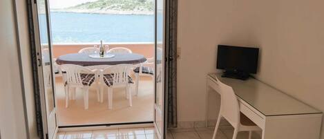 A room with a balcony, a table with chairs, a TV on a desk, and a view of the sea.