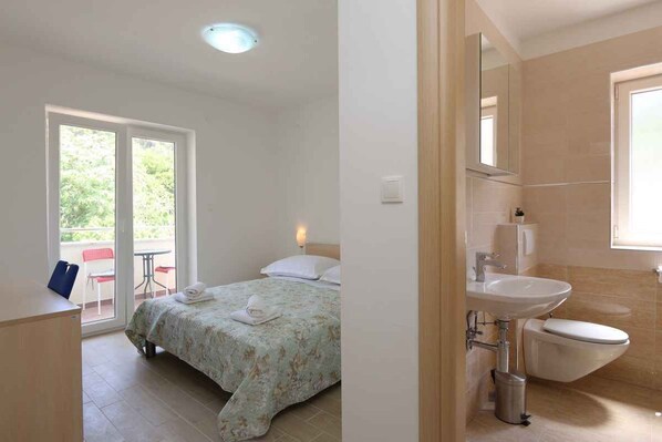 A bedroom with a bed, a bedside table, a window, and a bathroom with a sink and toilet.