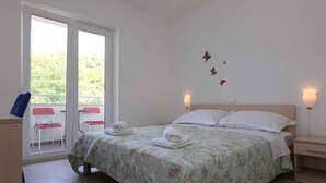 A bedroom with a bed, two bedside tables, a nightstand, a desk, two chairs, and a window with a view of greenery. - Holiday apartment with air conditioning only 40 m from the beach (Malinska)