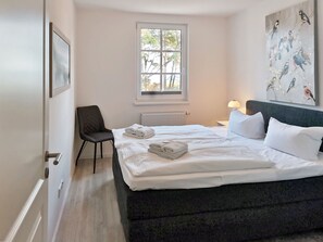 Un dormitorio con una cama, una silla, una ventana, un radiador y una lámpara fijada a la pared.
