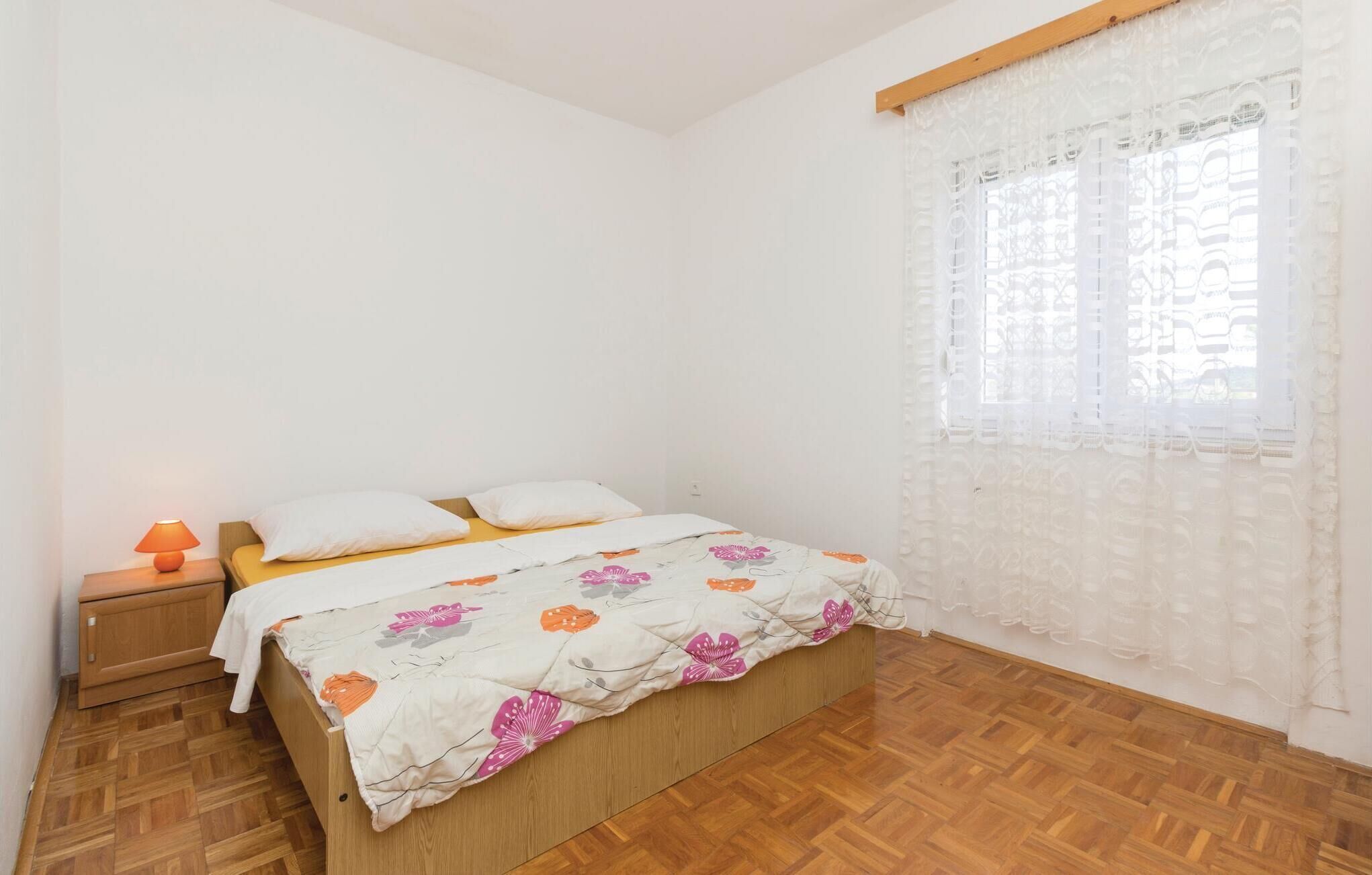 Un dormitorio con cama de madera, ropa de cama blanca, una mesita de noche con lámpara y una ventana con cortinas translúcidas.