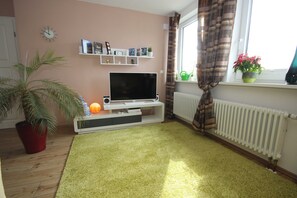 Uma sala de estar com televisão sobre um suporte, um vaso de planta, um radiador e uma janela com cortinas.