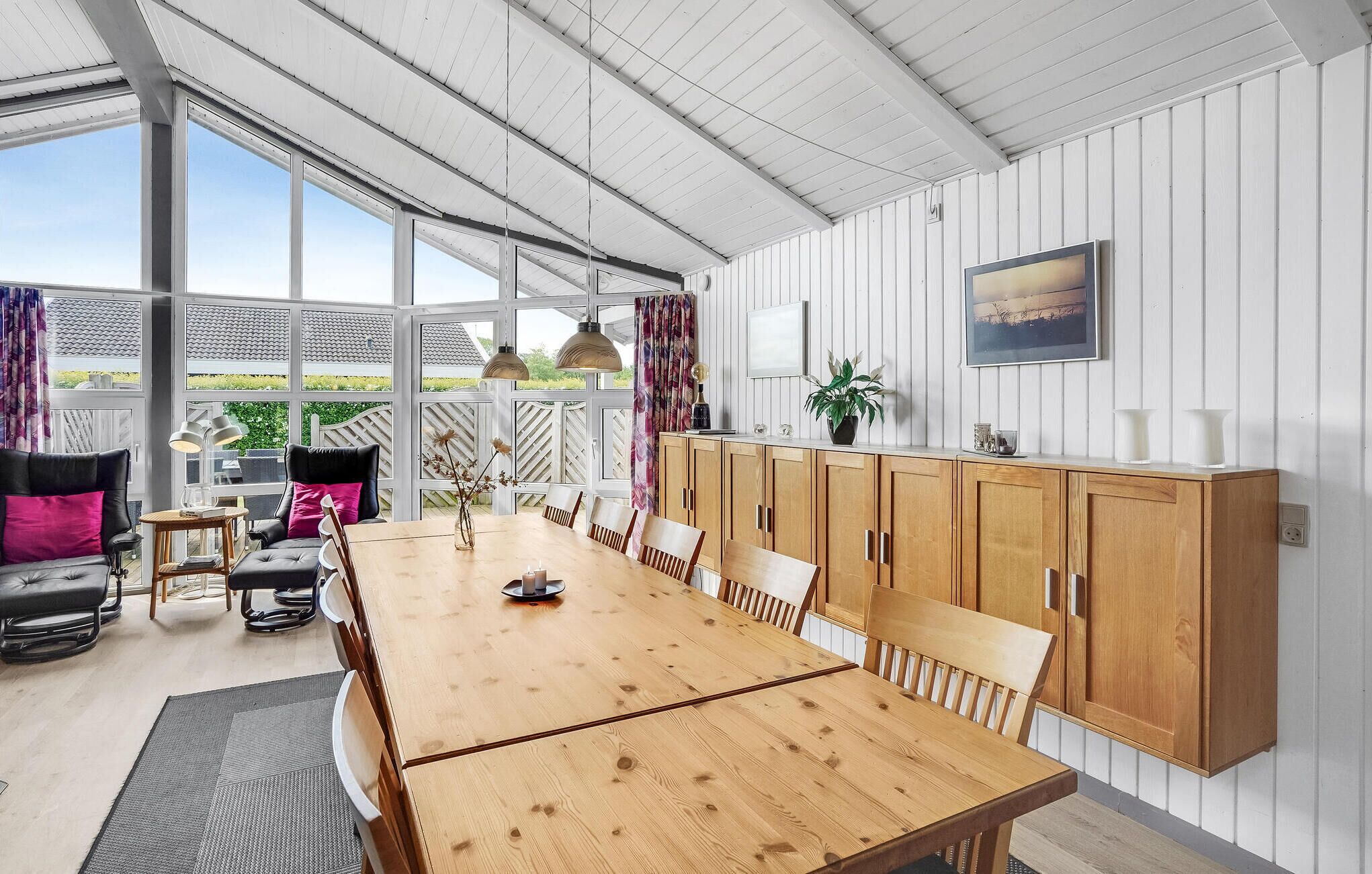 Un amplio comedor con una mesa y sillas de madera, un sofá y un armario. Grandes ventanales ofrecen vistas al exterior.