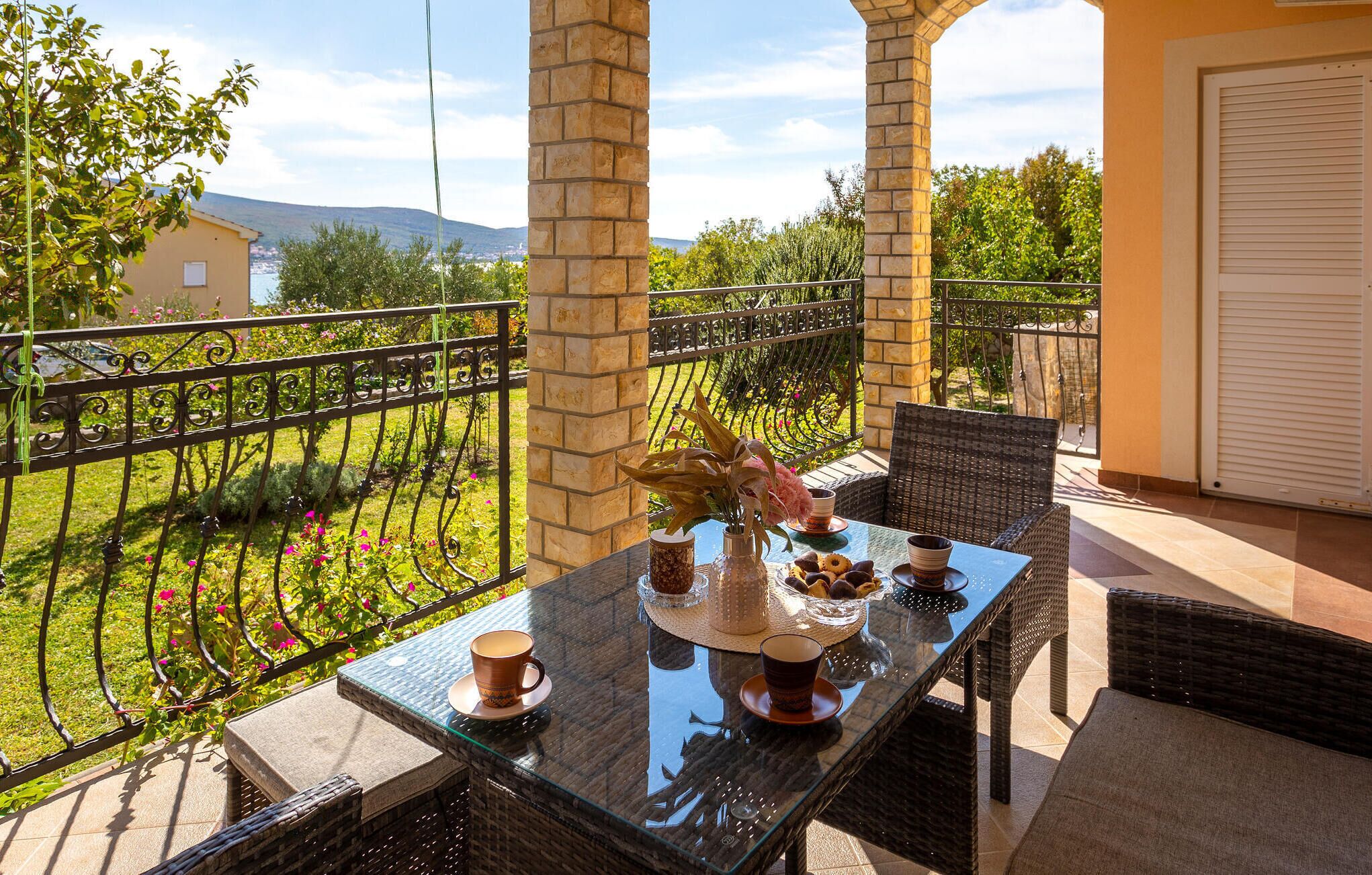 Ein Balkon mit Glastisch, Korbstühlen und Blick auf einen Garten sowie weit entfernte Hügel.