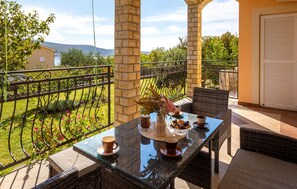 A balcony with a glass table, wicker chairs, and a view of a garden and distant hills. - Cozy apartment in Kornic with house sea view (Kornic)