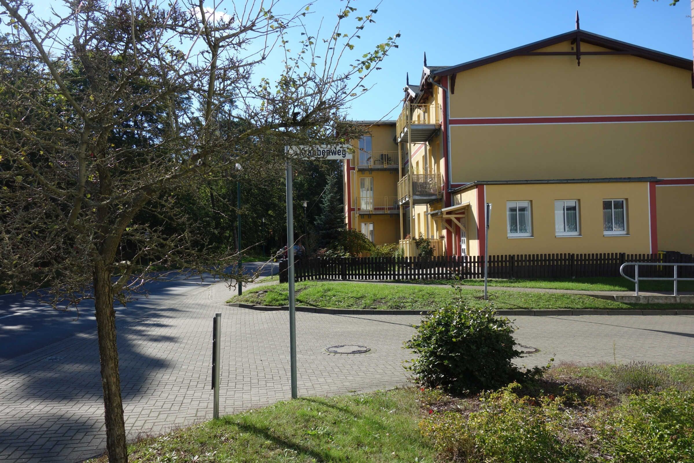 A building with a yellow facade, a black fence, and a paved area with a tree and bushes.