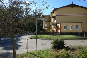 En byggnad med en gul fasad, ett svart staket och en asfalterad yta med ett träd och buskar.