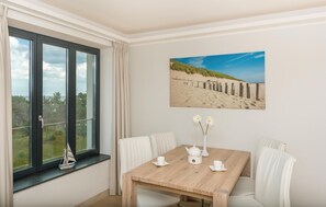 Een kamer met een groot raam, een houten tafel met witte stoelen, een schilderij van een strand en een klein model van een schip op de vensterbank.
