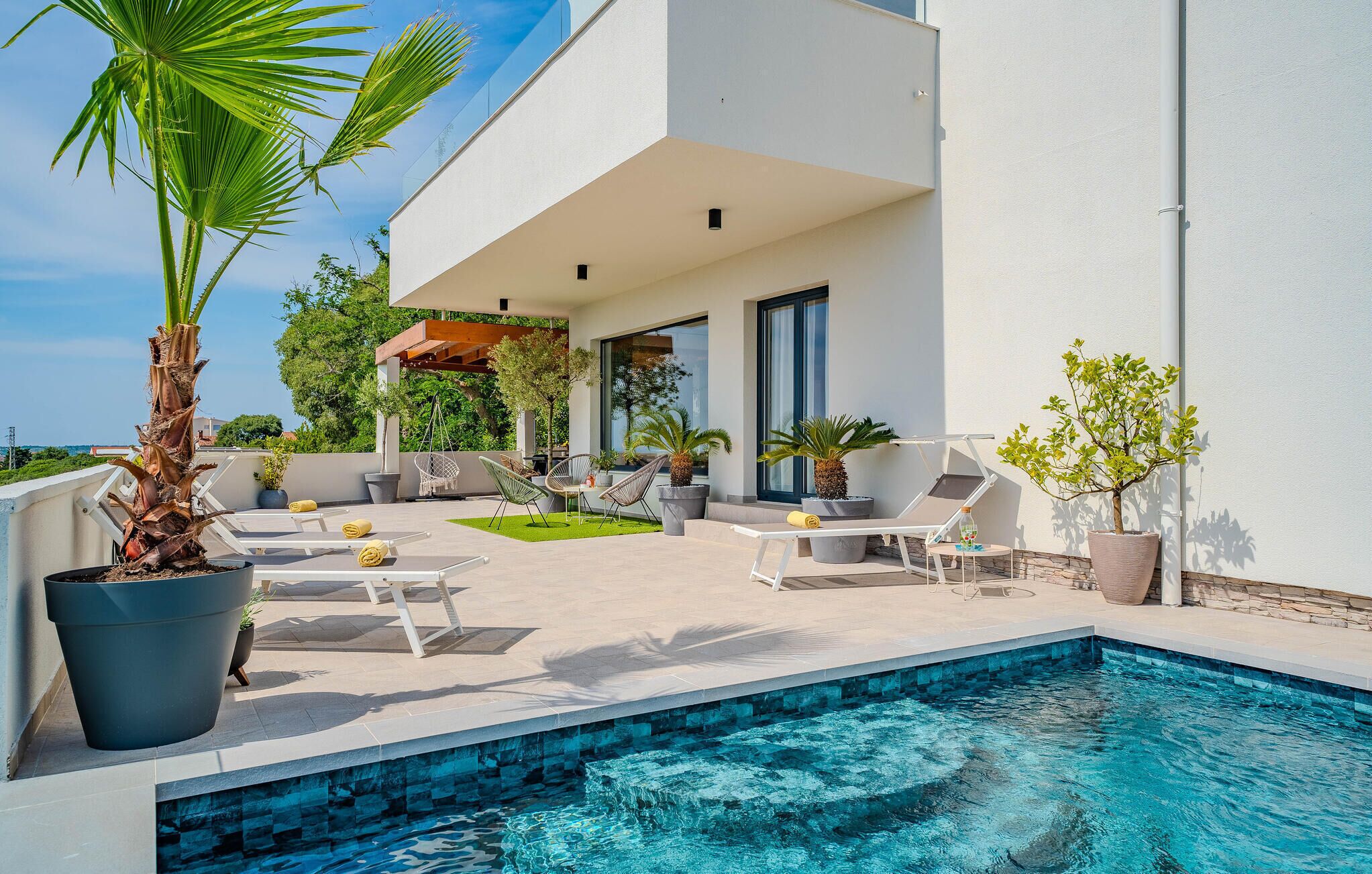 A modern outdoor pool area with lounge chairs, a hanging chair, and potted plants.