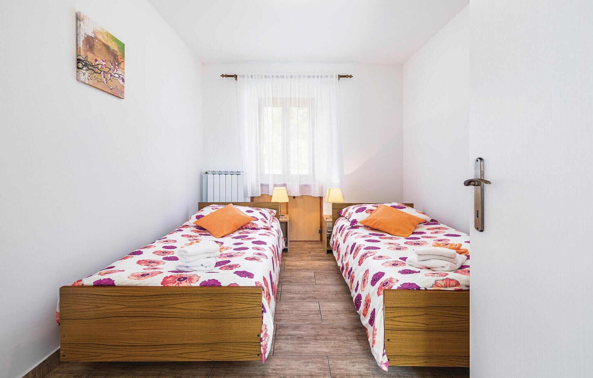A small, clean room with two wooden beds, white walls, a radiator, and a window with sheer curtains.