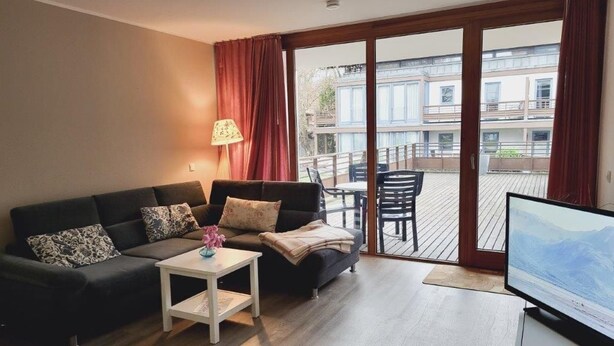 Un salon avec un canapé, une petite table avec une lampe et un téléviseur. Il y a un balcon avec vue sur les immeubles et une table à l’extérieur.