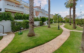 A well-maintained garden with palm trees, a stone pathway, and a white building with balconies. - Cozy apartment in Guardamar del Segura (Guardamar del Segura)