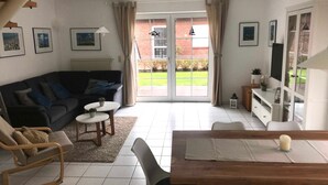 A living room with a sofa, coffee table, and a dining area with a table and chairs. There is a sliding glass door leading to a garden. - Vacation home in Nessmersiel 20-129a - 200-129a (Dornum)