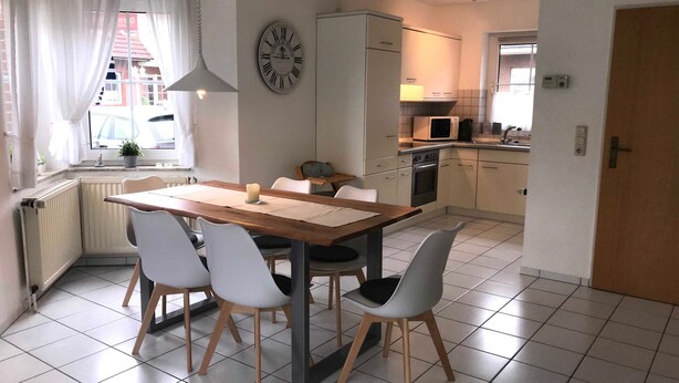 A modern kitchen with a dining area, featuring a wooden table, white chairs, a clock, and a microwave. - Vacation home in Nessmersiel 20-129a - 200-129a (Dornum)