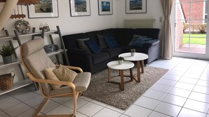 A living room with a black sofa, a wooden chair, a small white table, and a staircase. - Vacation home in Nessmersiel 20-129a - 200-129a (Dornum)