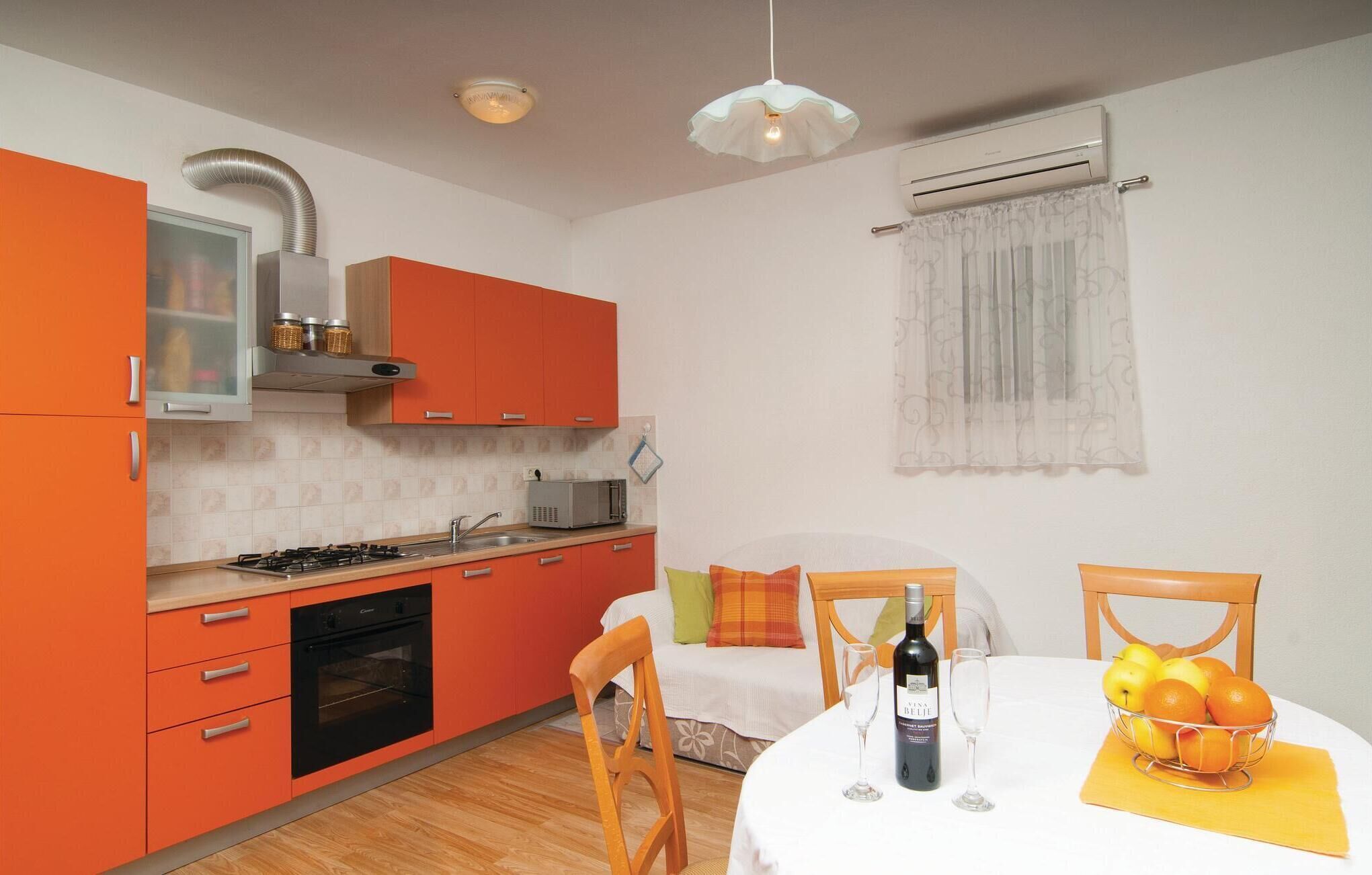 Een kleine keuken met oranje kastjes, een gasfornuis en een magnetron. Er is een eethoek met een tafel gedekt voor twee personen, een fles wijn en een fruitschaal.