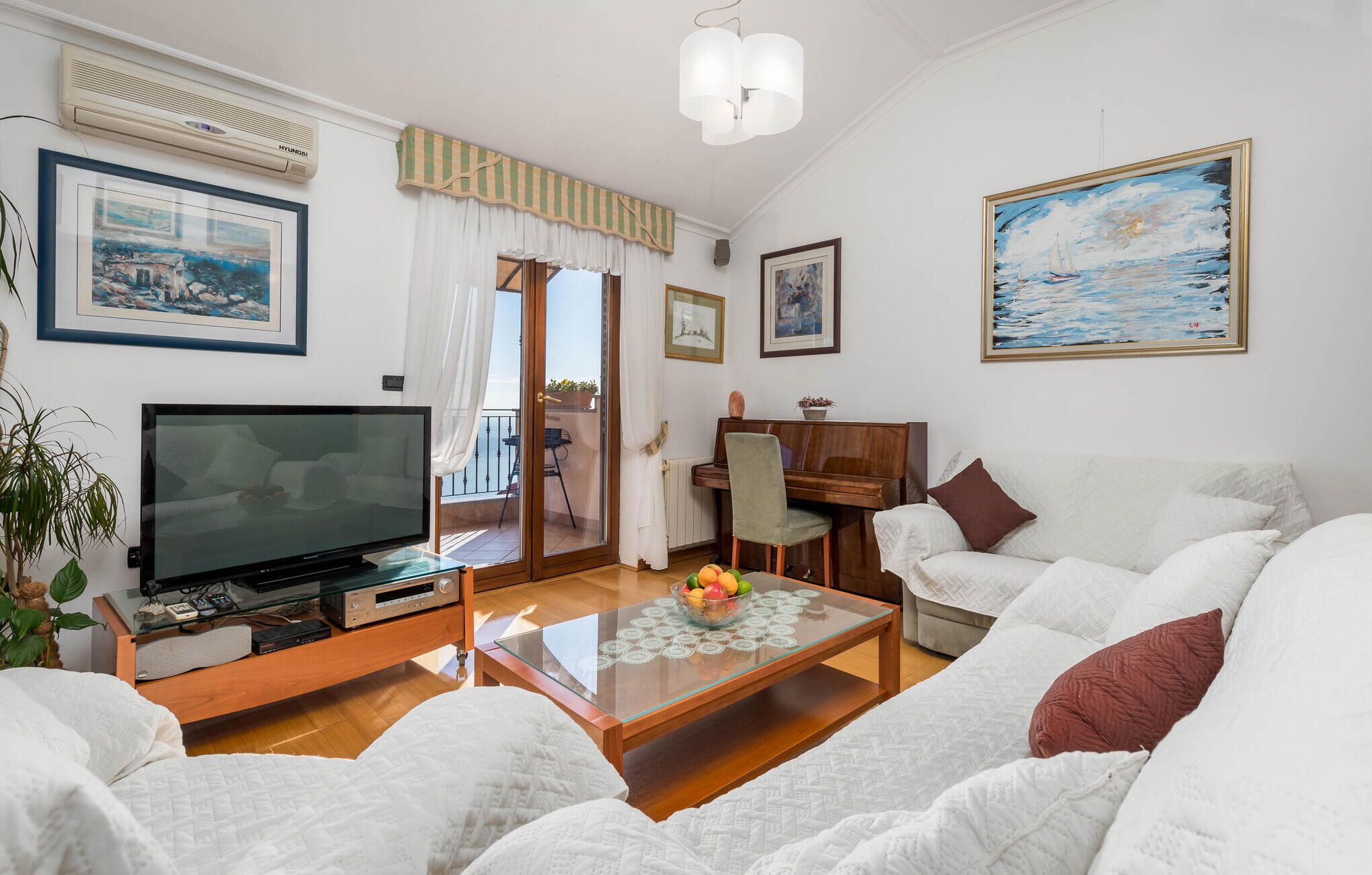 Ein Wohnzimmer mit Fernseher, Klavier, einem Glastisch mit Obst und einer Couch.