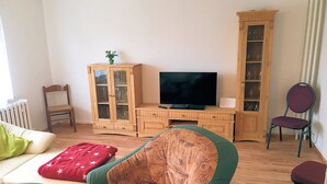 A living room with a sofa, a green armchair, a wooden cabinet with glass doors, a television, and a chair. - Wede, WH - Residential building, Germany (Ückeritz)