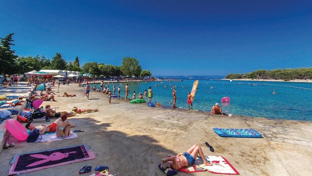 Een strand met mensen die zonnebaden en zwemmen, een heldere lucht en een kalme zee.