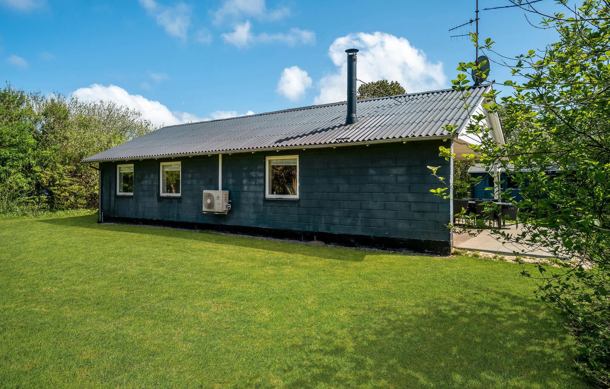 Une petite maison de plain-pied, à l’extérieur d’un bleu foncé, avec un toit en tuiles et une cheminée. Elle comporte trois fenêtres et un climatiseur fixé au mur.