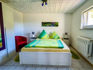 A bedroom with a bed, a red chair, a nightstand, a lamp, a radiator, and a window.
