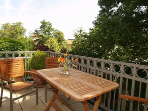 A wooden table and chairs on a balcony with a metal railing, surrounded by greenery.