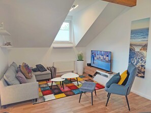 Ein modernes Loft-Apartment mit schrägem Dach, großem Fernseher, farbenfrohem Teppich und einem Strand-Gemälde.