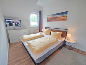 Ein Schlafzimmer mit Bett, Fernseher, Fenster, einem Gemälde und einem Nachttisch mit Lampe.