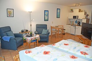 En kompakt opholdsstue med sofa, stol, lille spisebord, fjernsyn og tekøkken.