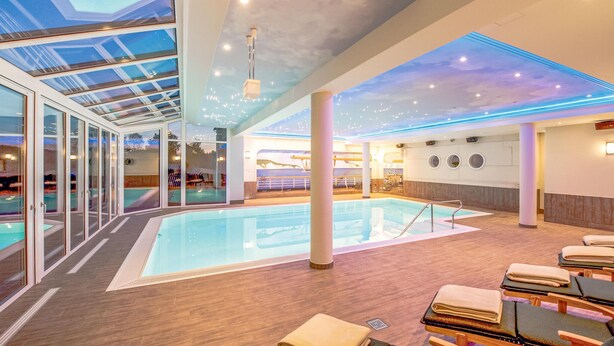 Ein moderner Poolbereich im Innenbereich mit Glaswänden, einem zentralen Schwimmbecken und Liegestühlen.