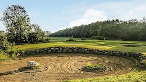 Ein Golfplatz mit Steinmauer, einem großen Baum und einem Wald im Hintergrund.