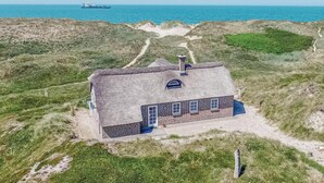 Een gebouw met rieten dak en een gevel van baksteen, gelegen op een grasduin met uitzicht op de zee.