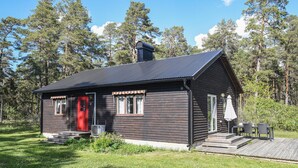 Een klein houten vakantiehuisje met een donkere houten gevel, een rode deur en een schoorsteen. Het heeft een klein terras met treden die naar de ingang leiden.