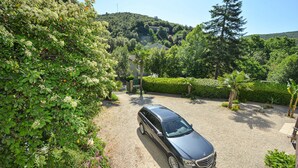 Une voiture noire est garée sur une allée de gravier, entourée d’une végétation luxuriante et avec une colline en arrière-plan.
