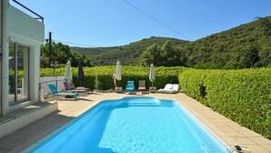 Une piscine entourée de chaises longues et de parasols, bordée d’une haie et avec des collines en arrière-plan.