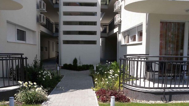 A pathway leading to a residential building with balconies, a small garden area, and a paved walkway. - Holiday apartment with pool Wi-Fi and tasteful decoration (Siófok)