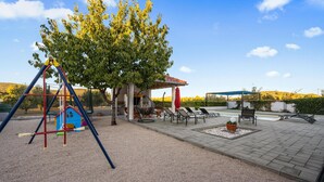 Ein Spielplatz mit Schaukeln und einem kleinen, blauen Spielhäuschen, einem überdachten Sitzbereich mit Stühlen und einem Tisch sowie ein Pool im Hintergrund.