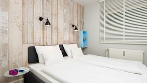 Un dormitorio moderno con pared de acento de madera, una cama con ropa de cama blanca, una mesita de noche pequeña con una lámpara y un mueble azul para guardar objetos.
