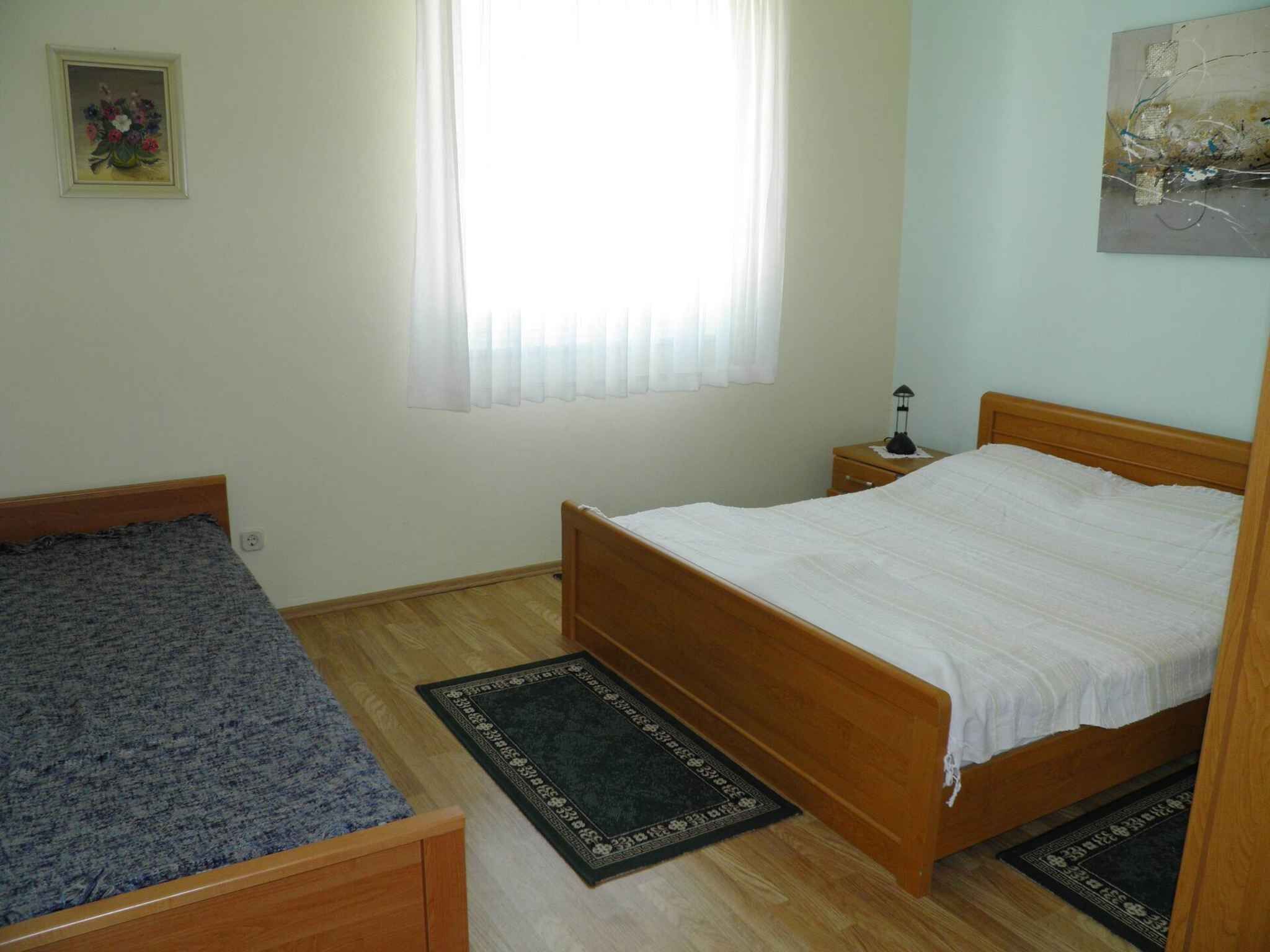 A bedroom with a single bed, a smaller bed, a nightstand, a lamp, and a window with sheer curtains.