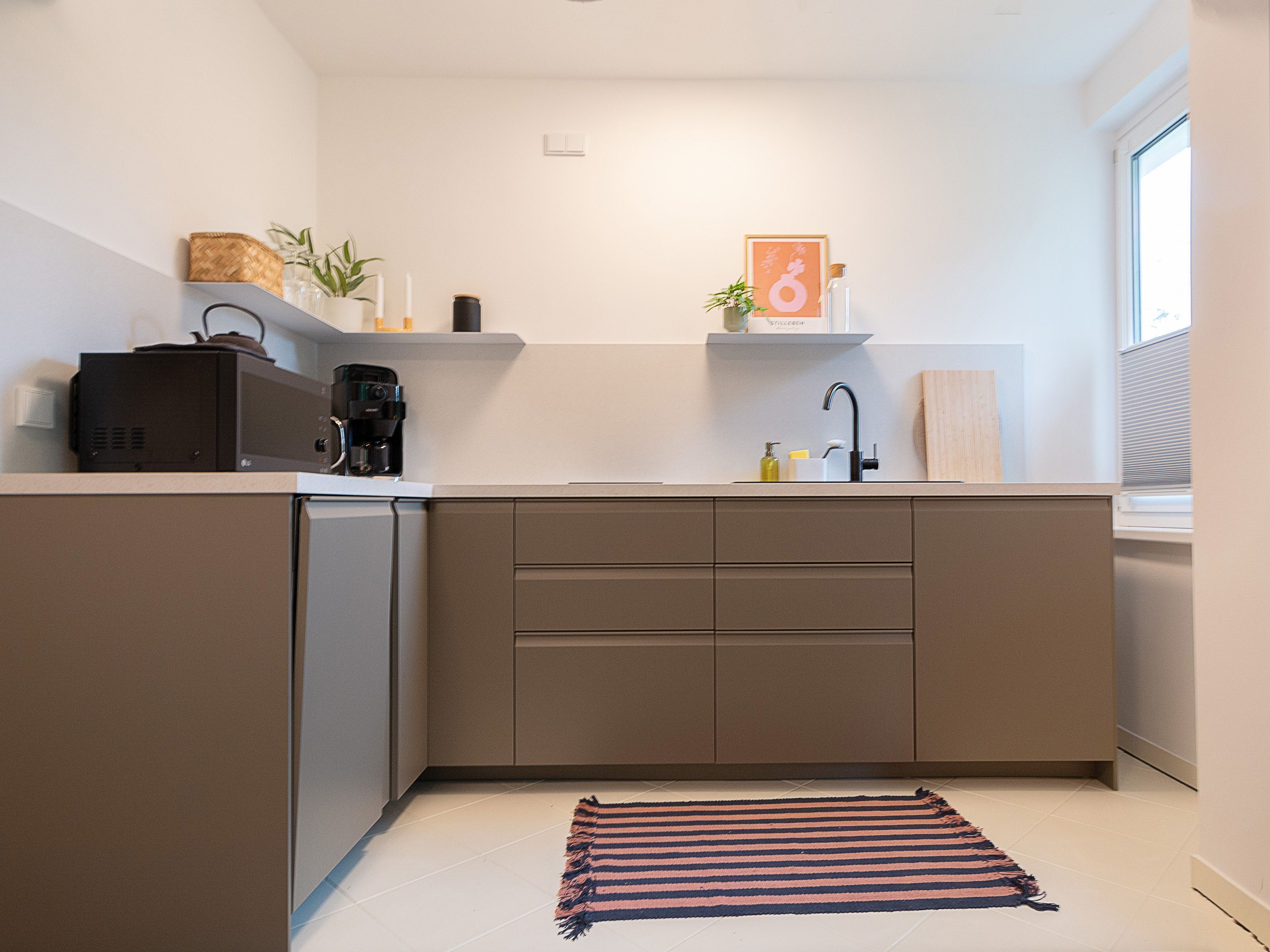 A modern kitchen with a built-in dishwasher, a coffee maker, and a sink with a faucet. There is a striped rug on the floor and a window on the right side.