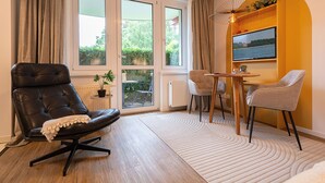 A modern living room with a leather chair, a dining table with chairs, a TV mounted on the wall, and large windows with curtains. - Dune garden Whg. Wa45-31 - Wa45-31 (Kühlungsborn)