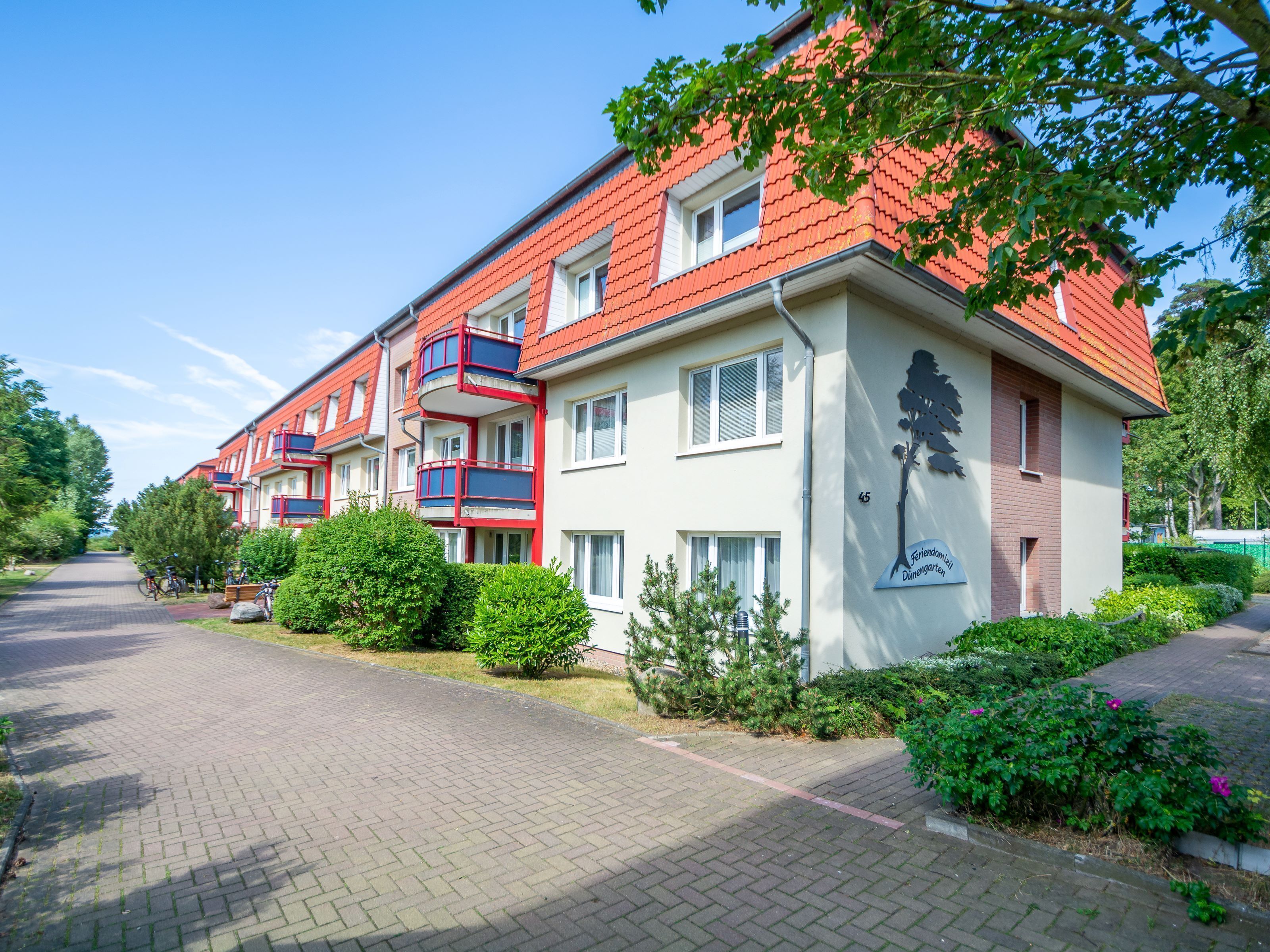 Une rangée d’immeubles d’appartements aux toits rouges, dotés de balcons, avec une allée pavée et une végétation bien entretenue.