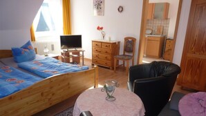 A bedroom with a bed, a desk, a chair, a small table with a glass, and a kitchenette in the background.