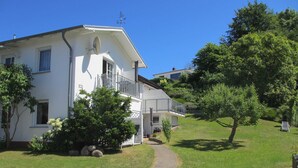 A two-story white building with a balcony, surrounded by greenery and a clear sky. - 1-room apartment on the ground floor with south-west facing terrace - Ferienhaus am Schmachter See (Binz)