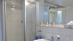 A bathroom with a glass shower enclosure, a white sink with a mirror above it, and a shelf with glass containers. - 1-room apartment on the ground floor with south-west facing terrace - Ferienhaus am Schmachter See (Binz)