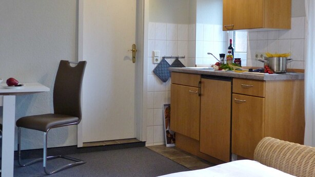 A compact kitchen with a dining area, a chair, and a bed with a woven headboard. - 1-room apartment on the ground floor with south-west facing terrace - Ferienhaus am Schmachter See (Binz)