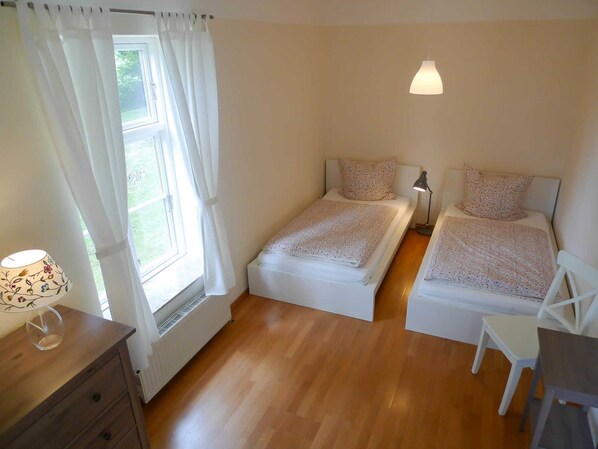 A bedroom with two single beds, a small desk, a chair, a lamp, and a window with curtains. - Apartment 1 - big apartments in the Manor House (Weitendorf)