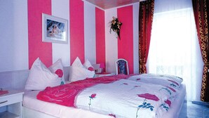 Een slaapkamer met een bed, kussens, een nachtkastje, een spiegel, een schilderij aan de muur en een raam met gordijnen.