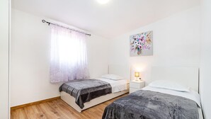 Un dormitorio con dos camas, una mesita de noche, una lámpara, una ventana con cortinas translúcidas y una obra de arte floral en la pared.